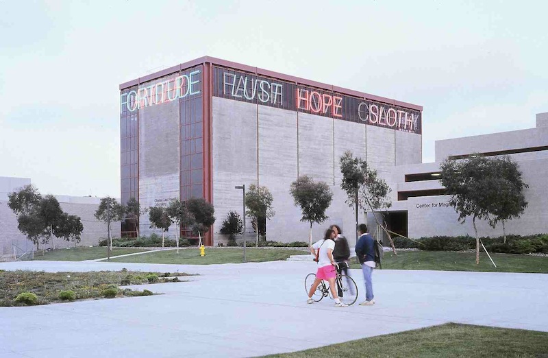 A photograph of a six-story concrete and glass building towering over the relatively smaller concrete classroom buildings of a university campus, surrounded by green sapling trees on neatly landscaped lawns, and wide pedestrian paths. A group of three people, one standing beside a bicycle occupies the foreground. On the top story of the otherwise undecorated tower, neon letters forming words for vices and virtues are arrayed in a frieze and flash in an alternating pattern. (Nauman, Bruce. 1988. Vices and Virtues. Neon tubing. Stuart Collection, University of California, San Diego. <https://stuartcollection.ucsd.edu/artist/nauman.html>.)