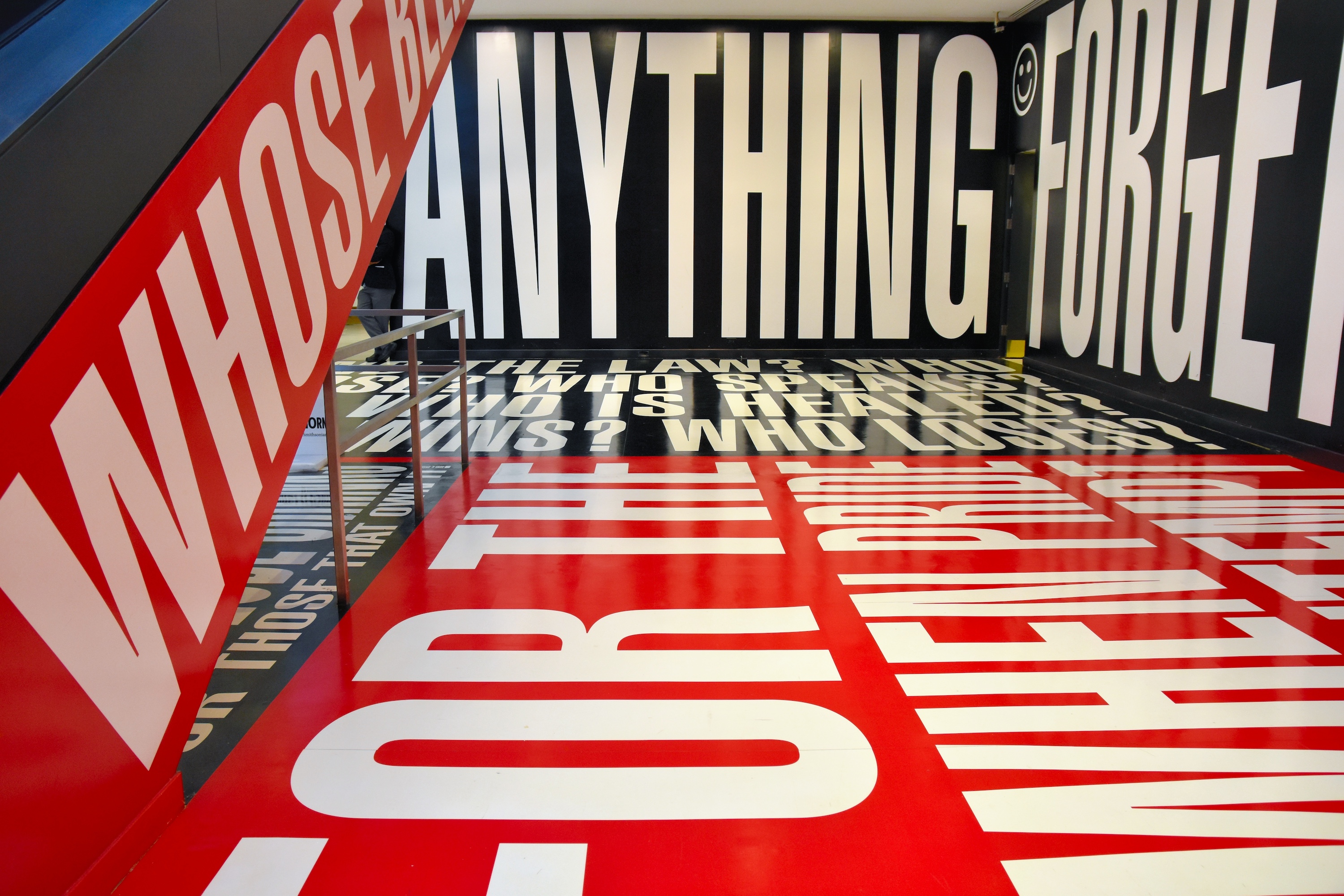 A photograph of an interior hallway framed by the side of an escalator on the left extending the next floor. The side of the escalator, the walls, ceiling, and floor, are covered with gleaming text-printed vinyl panels that fill the entire space and bearing words and phrases in bold, white block letters on alternating black and red backgrounds, creating an immersive experience in which one is assaulted by provocative questions, like “WHO IS SILENT?” (Kruger, Barbara. 2012. *Belief + Doubt [Photo by jpellgen (@1105_jp)]*. Chromogenic print, in artist’s frame. Hirshhorn Museum, Smithsonian Institute, District of Columbia. [https://www.flickr.com/photos/jpellgen/46955365515/](https://www.flickr.com/photos/jpellgen/46955365515/).)