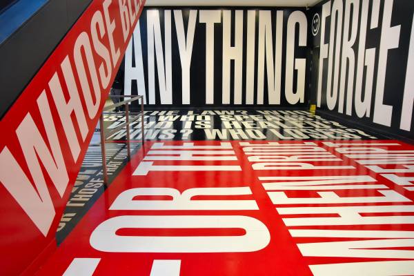 A photograph of an interior hallway framed by the side of an escalator on the left extending the next floor. The side of the escalator, the walls, ceiling, and floor, are covered with gleaming text-printed vinyl panels that fill the entire space and bearing words and phrases in bold, white block letters on alternating black and red backgrounds, creating an immersive experience in which one is assaulted by provocative questions, like “WHO IS SILENT?” (Kruger, Barbara. 2012. *Belief + Doubt [Photo by jpellgen (@1105_jp)]*. Chromogenic print, in artist’s frame. Hirshhorn Museum, Smithsonian Institute, District of Columbia. [https://www.flickr.com/photos/jpellgen/46955365515/](https://www.flickr.com/photos/jpellgen/46955365515/).)