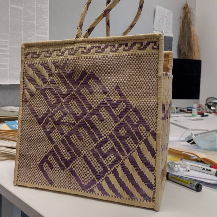 A basket made from finely woven material in natural color and purple dyed fibers
with square sides. The purple fibers are woven in a pattern that spells out...
