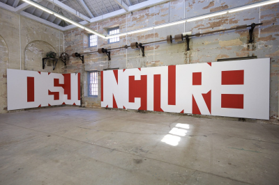 Wall partitions bearing large block letters in white on a dark red background sit against the interior walls of a hollowed out industrial warehouse (repurposed as an exhibition space), rising from floor to approximately 2 meters and wrapping around a corner. The letters are adjacent, leaving only the voids of the letterforms as negative space, and creating an uncanny effect while still being legible as a word, 'DISJUNCTURE.' (Nolan, Rose. 2019. A Big Word – DISJUNCTURE [Photo by Ian Hobbs]. Acrylic on wall partitions. <https://annaschwartzgallery.com/artist/rose-nolan>.)