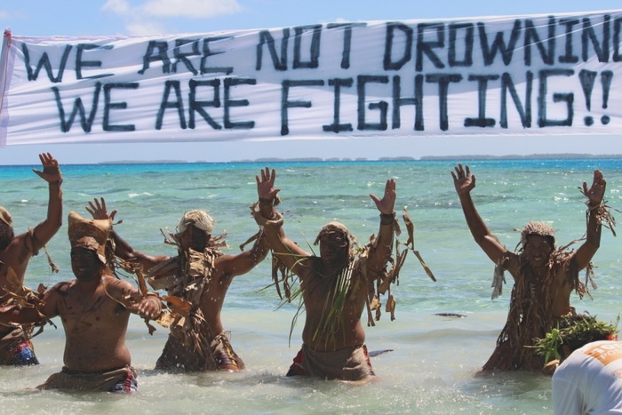 Men wearing Polynesian decorations and shorts stand waist deep in surf with hands raised under a banner reading “We are not drowning! We are fighting!!”